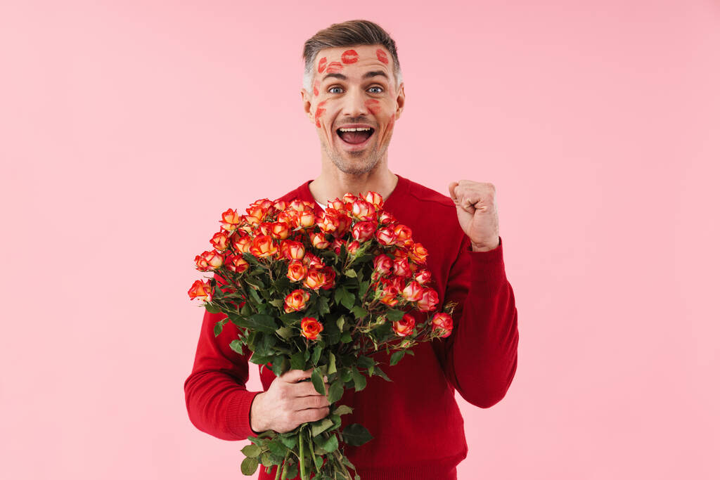 Retrato de belo homem caucasiano com marcas de beijo em seu rosto segurando flores no dia dos namorados isolado sobre fundo rosa
 - Foto, Imagem