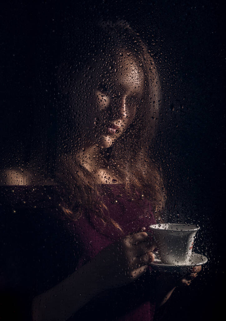 ωραίο νεαρό κορίτσι πίνει καφέ πίσω από το υγρό γυαλί, σταγόνες βροχής στο γυαλί - Φωτογραφία, εικόνα