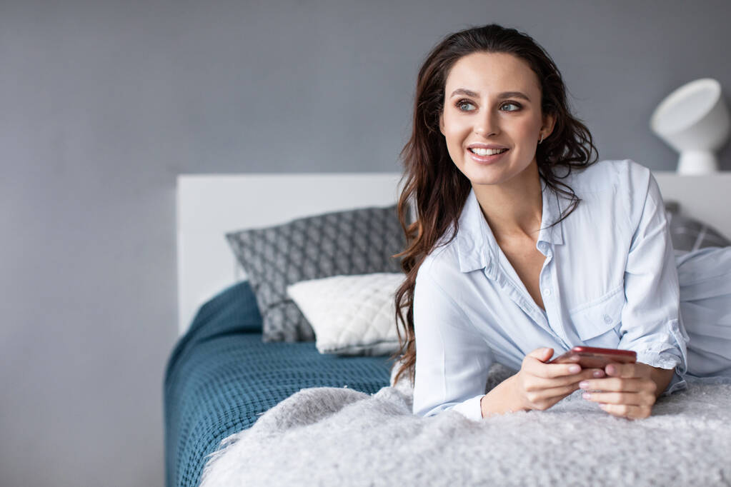 Retrato de bela mulher sorridente com telefone na cama. Use a internet móvel em casa
. - Foto, Imagem