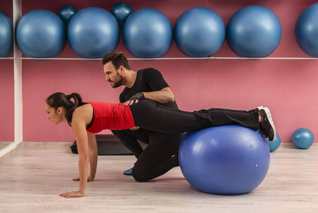 Mujer joven ayudado por su entrenador está haciendo ejercicios con una pelota de fitness en un gimnasio
. - Foto, Imagen