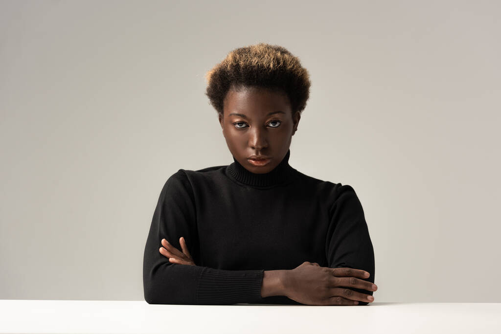 mujer afro en cuello alto negro sentada a la mesa con los brazos cruzados aislados en gris
 - Foto, imagen