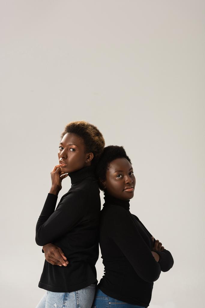 σκεπτόμενες Αφροαμερικανίδες με μαύρα ζιβάγκο να στέκονται πλάτη με πλάτη απομονωμένες σε γκρι. - Φωτογραφία, εικόνα