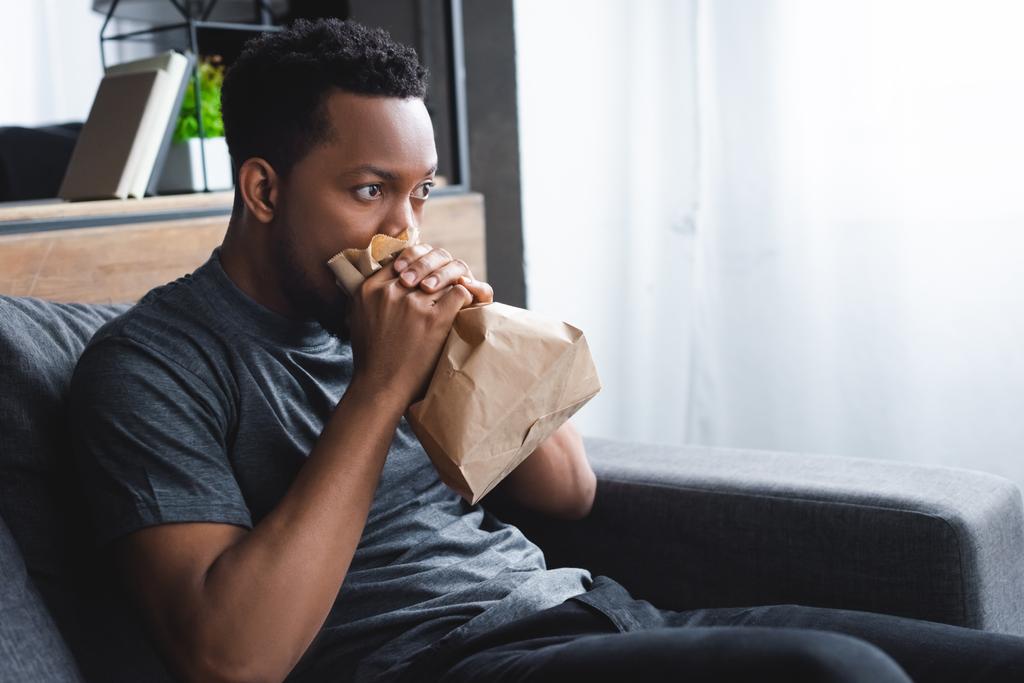 hombre afroamericano estresado respirando con bolsa de papel mientras tiene ataque de pánico en casa
 - Foto, Imagen