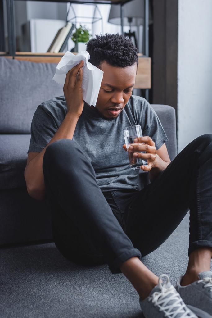 stressed sweaty african american man holding glass of water and napkins at home - Photo, Image
