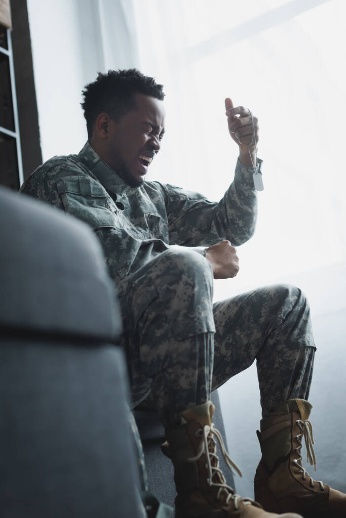 gestresster afrikanisch-amerikanischer Soldat schreit und hält Armeeabzeichen zu Hause, leidet an PTBS - Foto, Bild