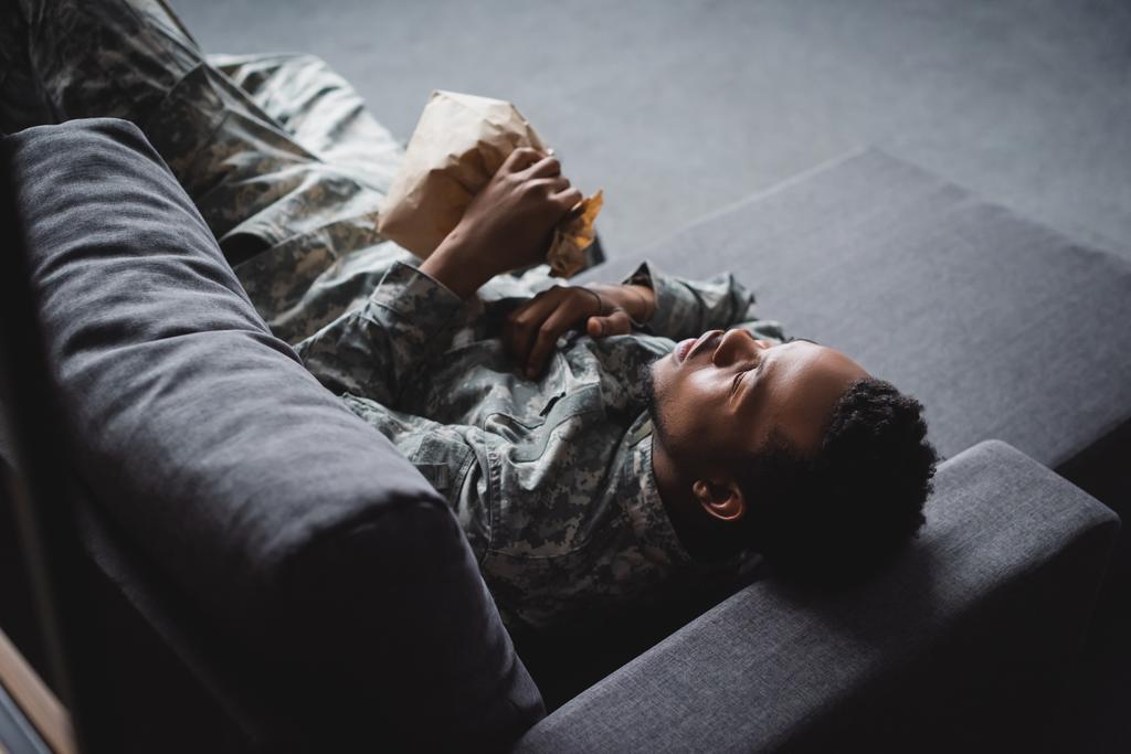 Afrikanischer amerikanischer Soldat in Militäruniform mit Papiertüte, während er zu Hause unter Panikattacken und PTBS leidet - Foto, Bild
