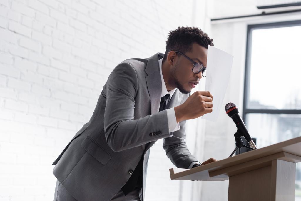 benadrukte Afrikaans Amerikaans spreker verbergen achter papier tijdens zakelijke conferentie - Foto, afbeelding