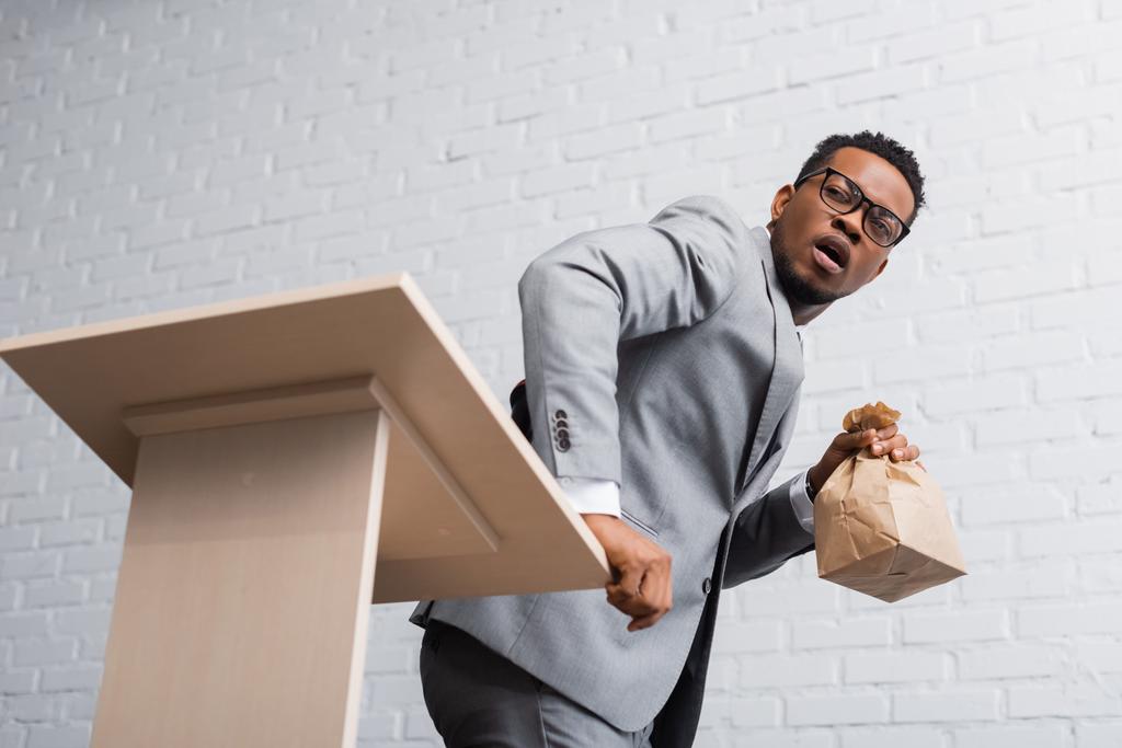 portavoz afroamericano asustado sosteniendo bolsa de papel y teniendo ataque de pánico durante conferencia de negocios en la oficina
  - Foto, Imagen
