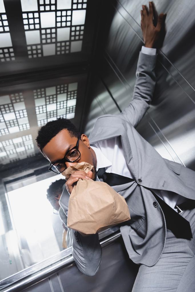 Afrika kökenli Amerikalı işadamı kağıt torbayla nefes alıyor ve asansörde panik atak geçiriyor. - Fotoğraf, Görsel