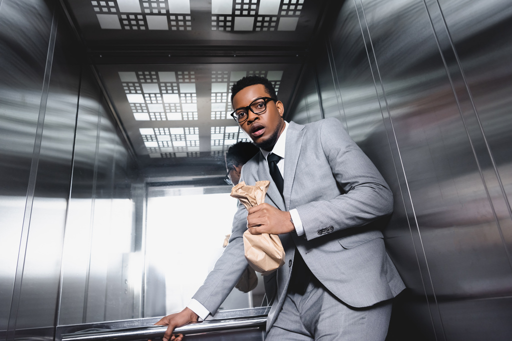 nervoso uomo d'affari afroamericano con sacchetto di carta che soffre di attacco di panico in ascensore
 - Foto, immagini