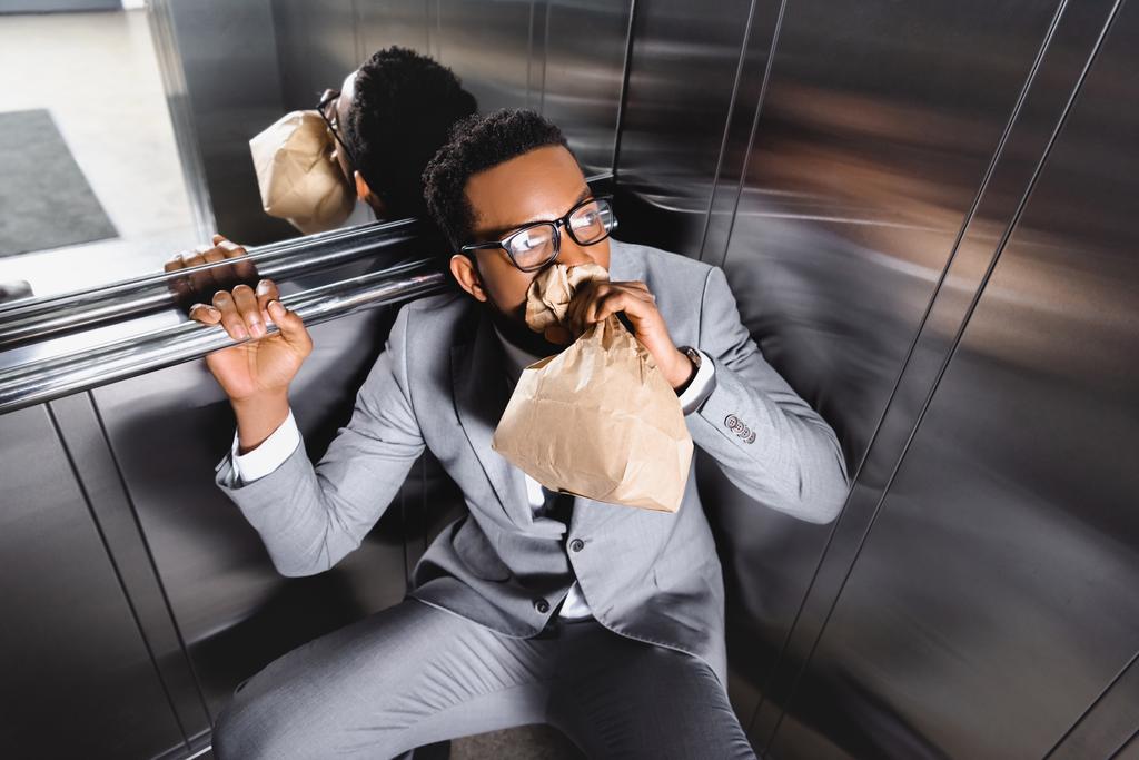 rémült afro-amerikai üzletember légzés papírzacskóval és szenved pánikroham a liftben - Fotó, kép