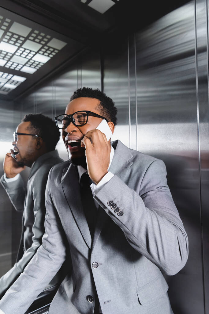 stressed african american businessman crying and talking on smartphone while suffering from panic attack in elevator - Photo, Image