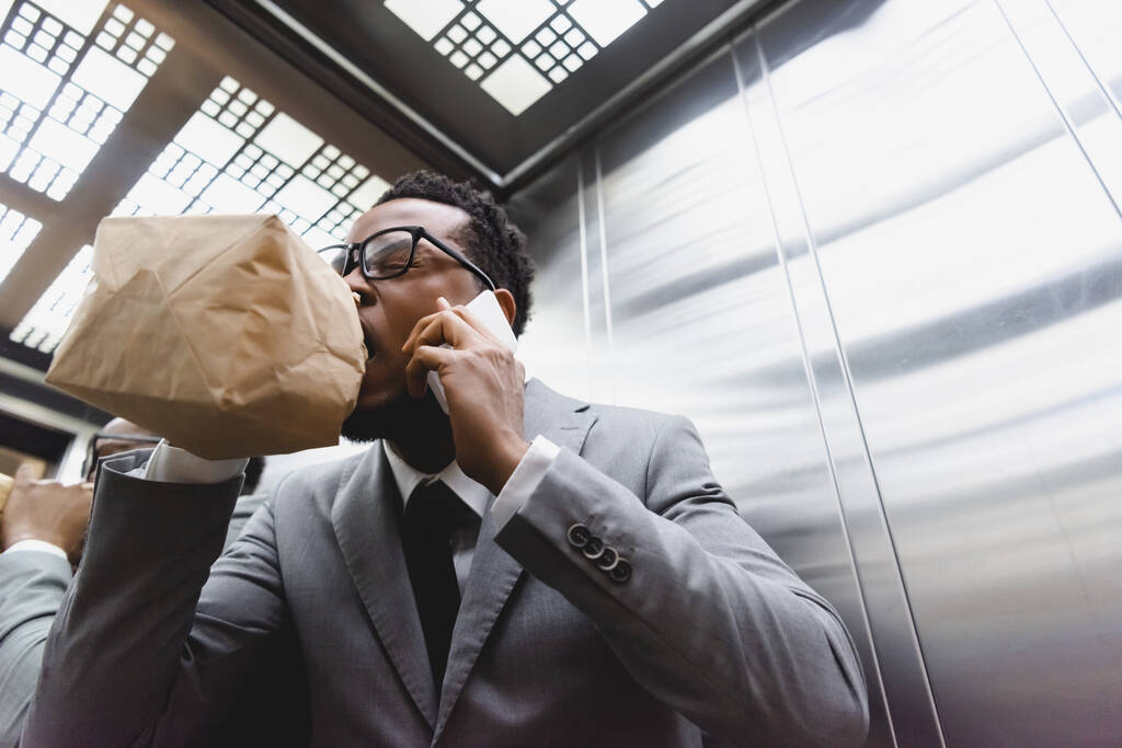 souligné homme d'affaires afro-américain parlant sur smartphone tout en respirant avec un sac en papier et souffrant d'une attaque de panique dans l'ascenseur
 - Photo, image
