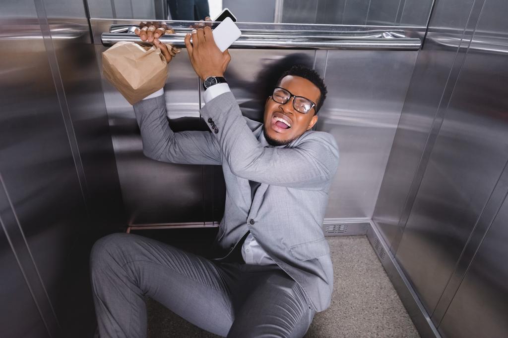 homem de negócios afro-americano estressado com smartphone e saco de papel sofrendo de ataque de pânico no elevador
 - Foto, Imagem