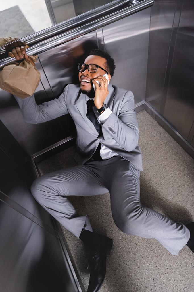 pláč africký americký podnikatel s papírovou taškou mluví na smartphone a trpí záchvat paniky ve výtahu - Fotografie, Obrázek