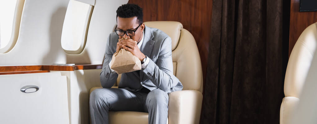panoramic shot of stressed african american businessman breathing with paper bag while having panic attack during flight on private plane - Photo, Image