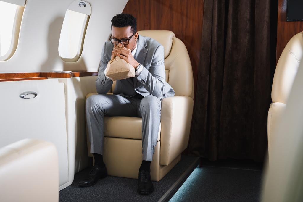 nervöser afrikanisch-amerikanischer Geschäftsmann atmet mit Papiertüte, während er während des Fluges im Privatflugzeug eine Panikattacke hat - Foto, Bild