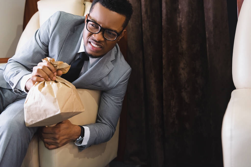 estresado empresario afroamericano sosteniendo bolsa de papel mientras tiene ataque de pánico durante el vuelo en avión
 - Foto, Imagen