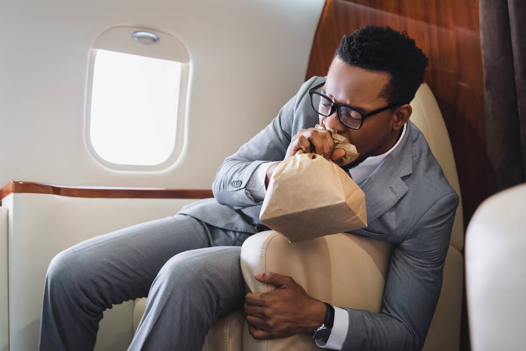 民間機での飛行中にパニック発作を起こしながらアフリカ系アメリカ人のビジネスマンが紙袋で呼吸する - 写真・画像