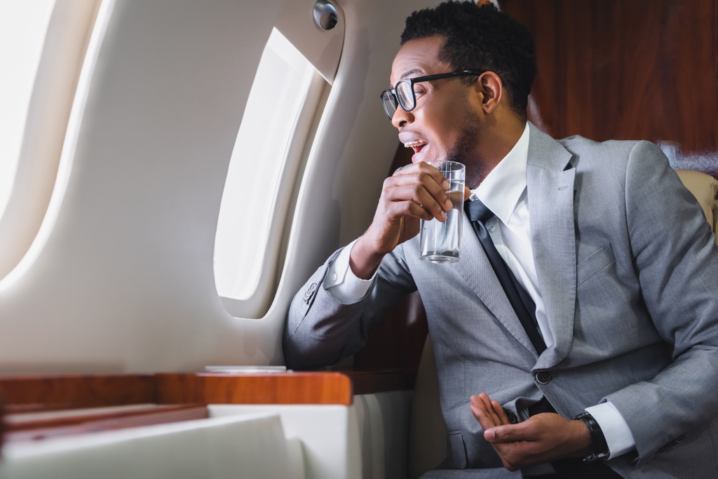 hombre de negocios afroamericano acentuado que mira a través de la ventana mientras sostiene las píldoras y el vaso de agua, sufriendo de ataque de pánico durante el vuelo en avión privado
 - Foto, imagen