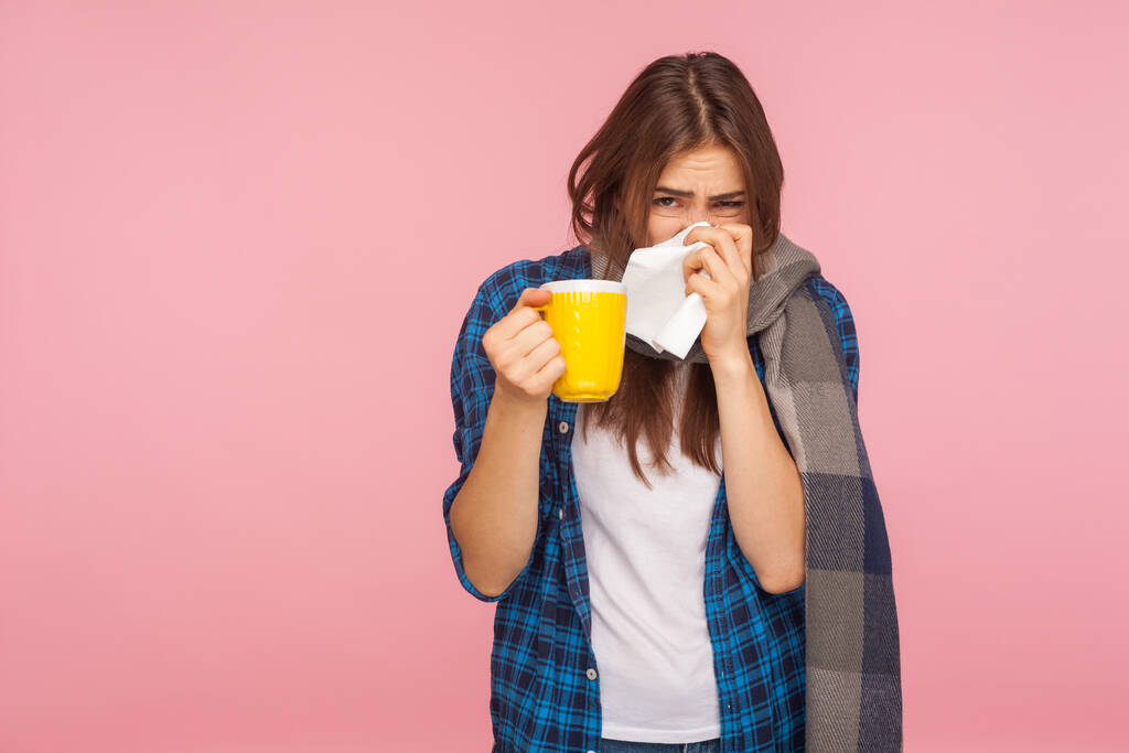 Лікування сезонного грипу. Хвора дівчина, загорнута в шарф, дме нежить, чхає в серветку і тримає чашку чаю, лікує грип, кашель і гарячку. критий студійний знімок ізольований на рожевому фоні
 - Фото, зображення