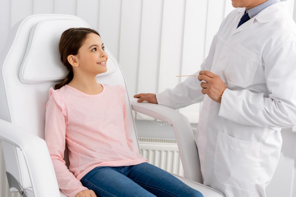 обрезанный вид отоларинголога, держащего лопатку в горле, стоя рядом с улыбающимся ребенком, сидящим в медицинском кресле
 - Фото, изображение