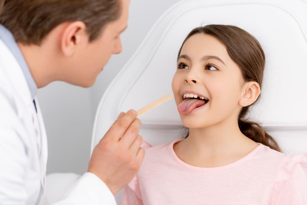 enfoque selectivo de ent médico sosteniendo depresor lengua cerca lindo niño sobresaliendo lengua
 - Foto, imagen
