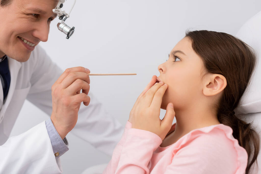 Lächelnder Arzt hält Zungendepressor in der Nähe verängstigtes Kind - Foto, Bild