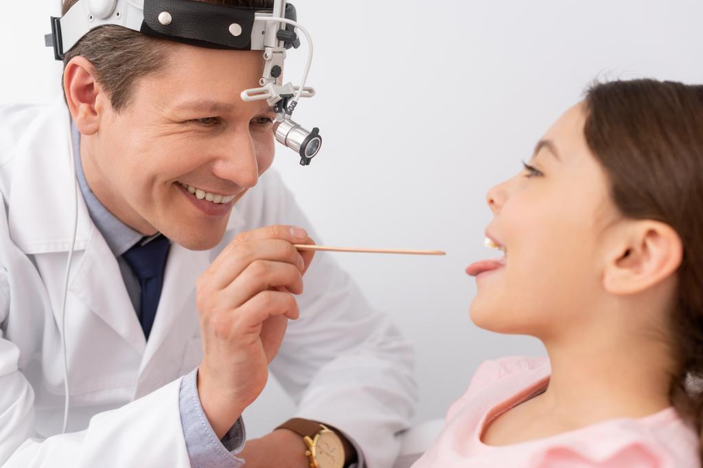 cheerful ent physician examining throat of cute child with tongue depressor - Photo, Image