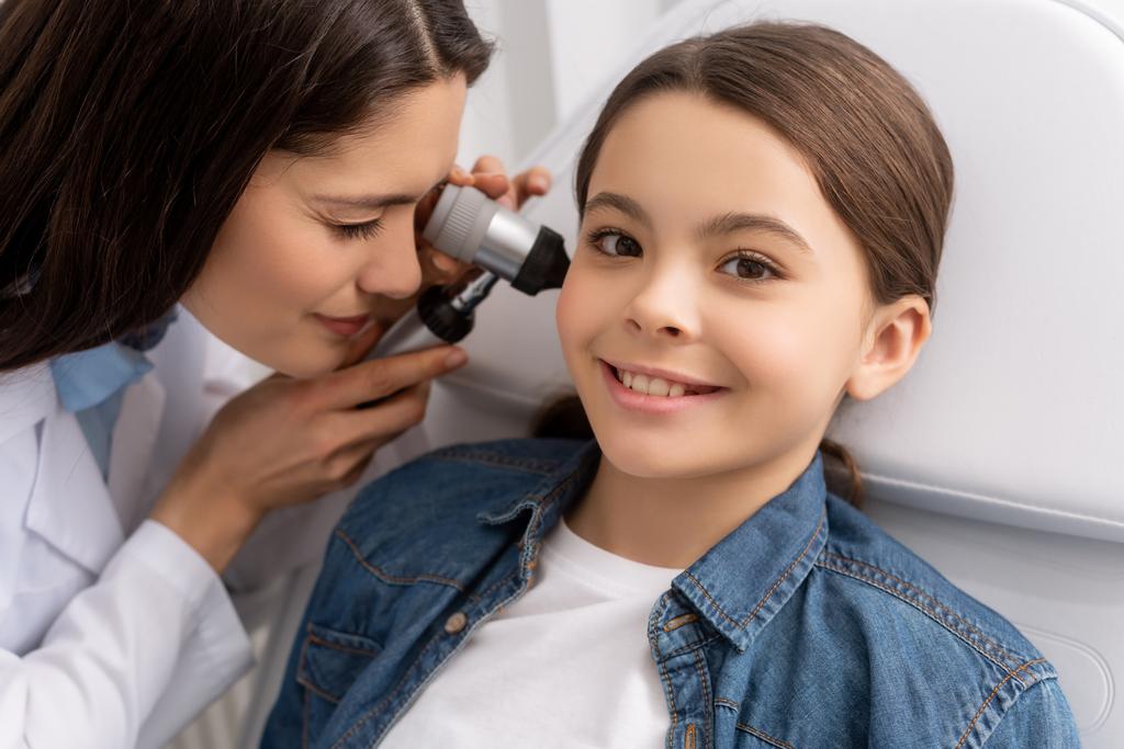 περιποιητικός ωτορινολαρυγγολόγος εξετάζει το αυτί του χαμογελαστού παιδιού με ωτοσκόπιο  - Φωτογραφία, εικόνα