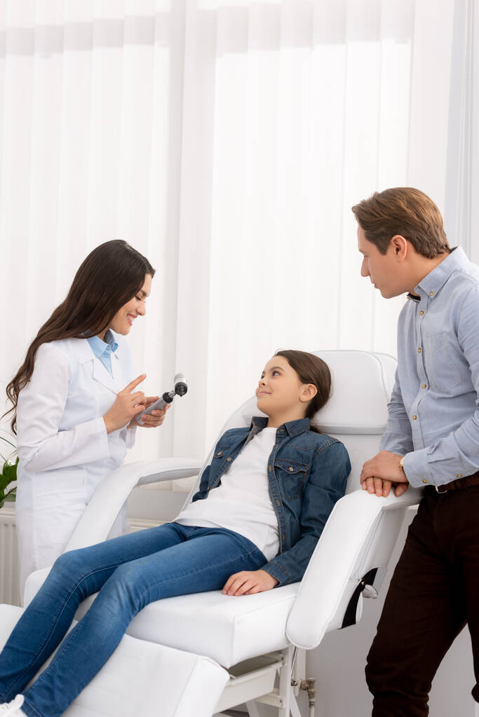отоларинголог показывает отоскоп улыбающемуся ребенку, сидящему в медицинском кресле, и ее отцу, стоящему рядом
 - Фото, изображение