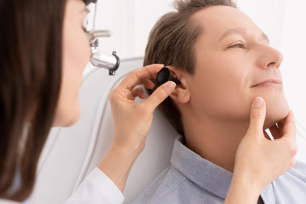 bijgesneden weergave van otolaryngoloog in ent koplamp onderzoeken oor van knappe patiënt  - Foto, afbeelding
