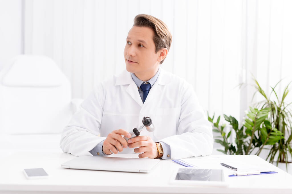 pohledný lékař drží otoskop a dívá se stranou, zatímco sedí na pracovišti - Fotografie, Obrázek
