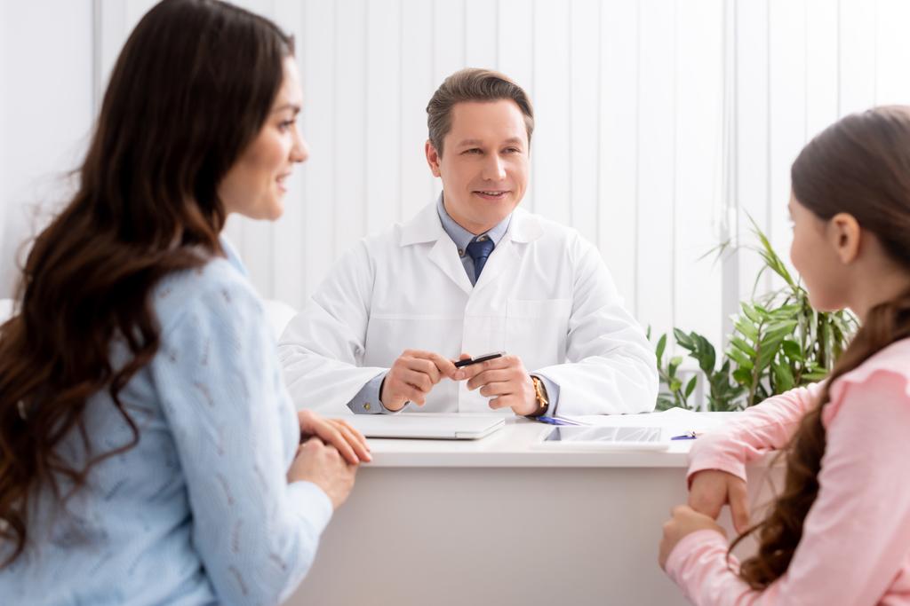 foco seletivo de mãe e filha sentadas perto de um médico sorridente durante a consulta
 - Foto, Imagem