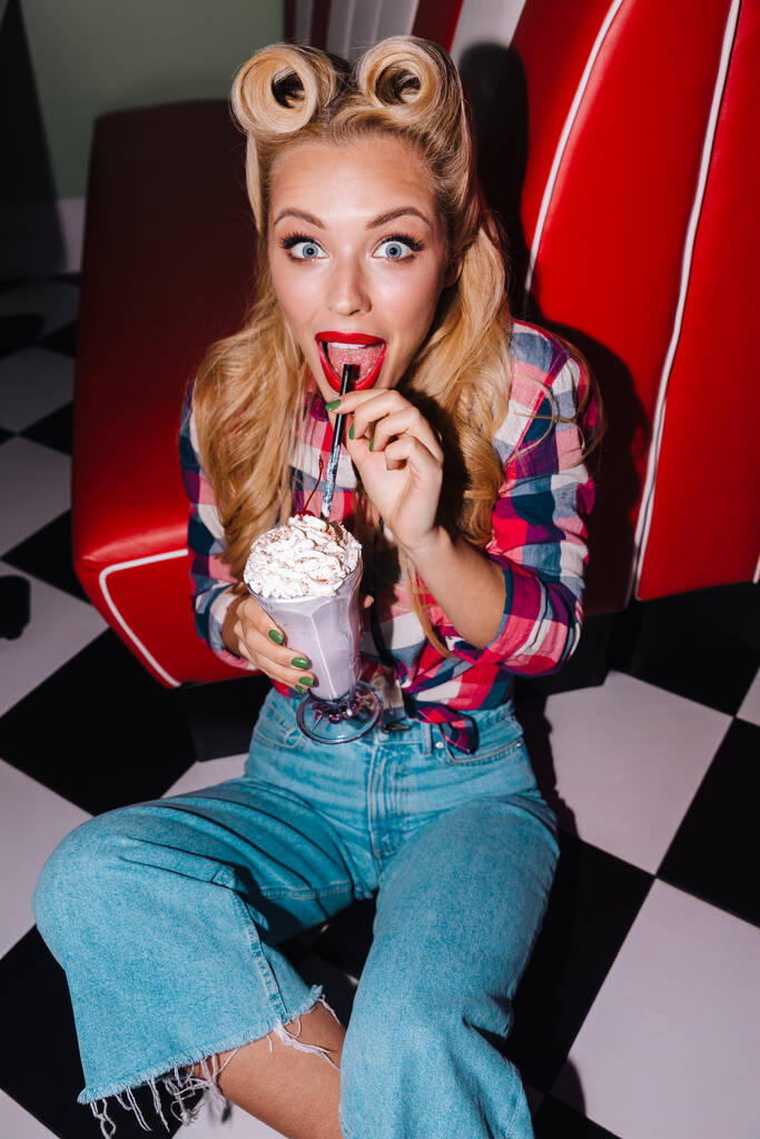 Foto de mulher alegre lindo com belo penteado bebendo milkshake enquanto sentado no chão no café retro americano
 - Foto, Imagem