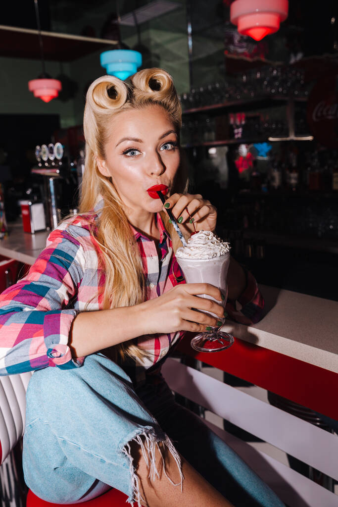 Foto de hermosa mujer alegre con hermoso peinado bebiendo batido en la cafetería retro americana
 - Foto, Imagen