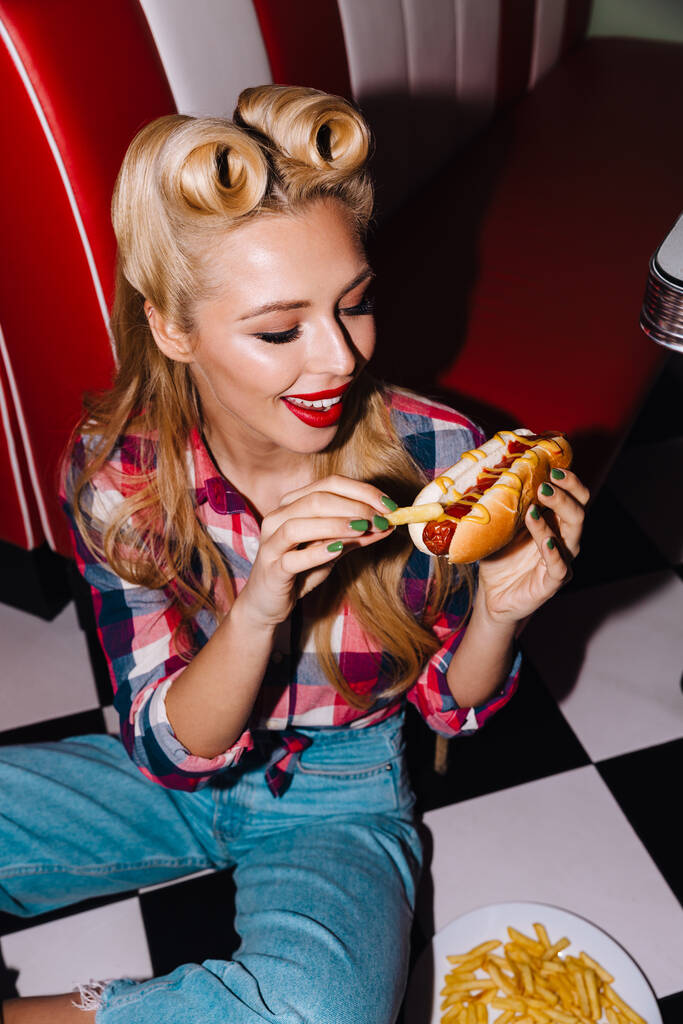 Φωτογραφία της νεαρής χαρούμενης γυναίκας με όμορφο χτένισμα τρώγοντας χοτ ντογκ και τηγανητές πατάτες σε ρετρό αμερικανική καφετέρια - Φωτογραφία, εικόνα