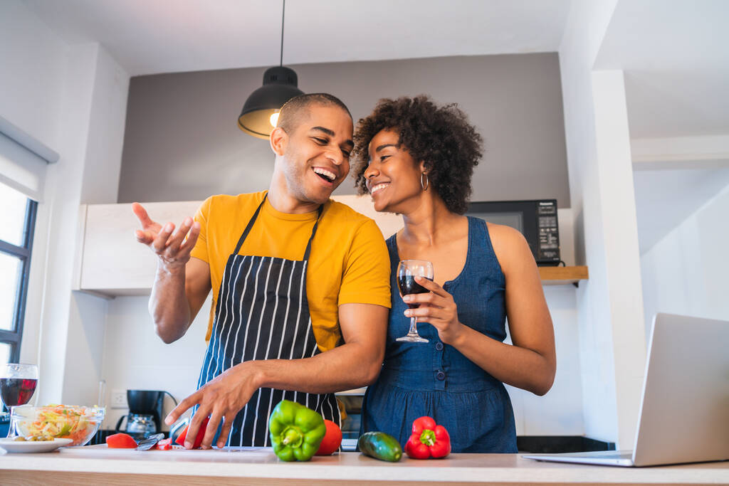 Porträt eines jungen lateinamerikanischen Paares, das einen Laptop benutzt, während es zu Hause in der Küche kocht. Beziehung, Koch und Lifestylekonzept. - Foto, Bild