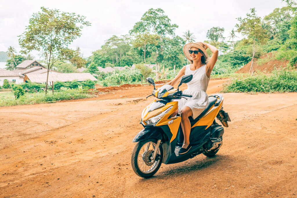 Beyaz elbiseli ve güneş gözlüklü mutlu kadın palmiye ağacının altında motosiklet sürüyor. Dikkatsiz tatil konsepti. - Fotoğraf, Görsel