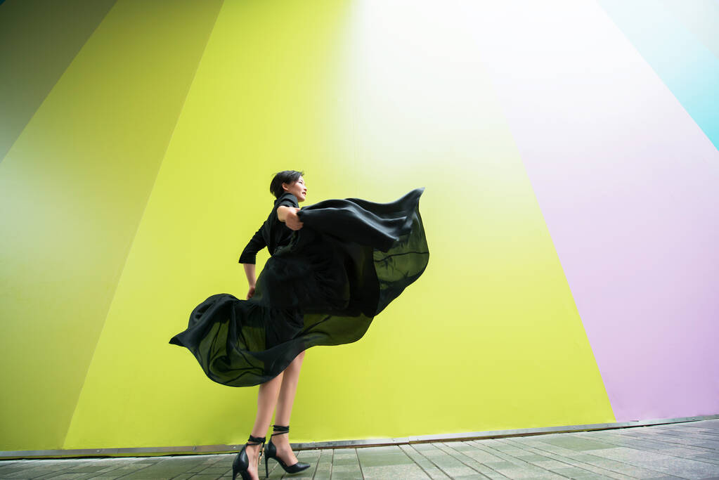 Donna di moda. Giovane bella ragazza cinese danza all'aperto indossando lungo abito nero con tacchi alti su sfondo muro colorato. Elegante signora alla moda.  - Foto, immagini