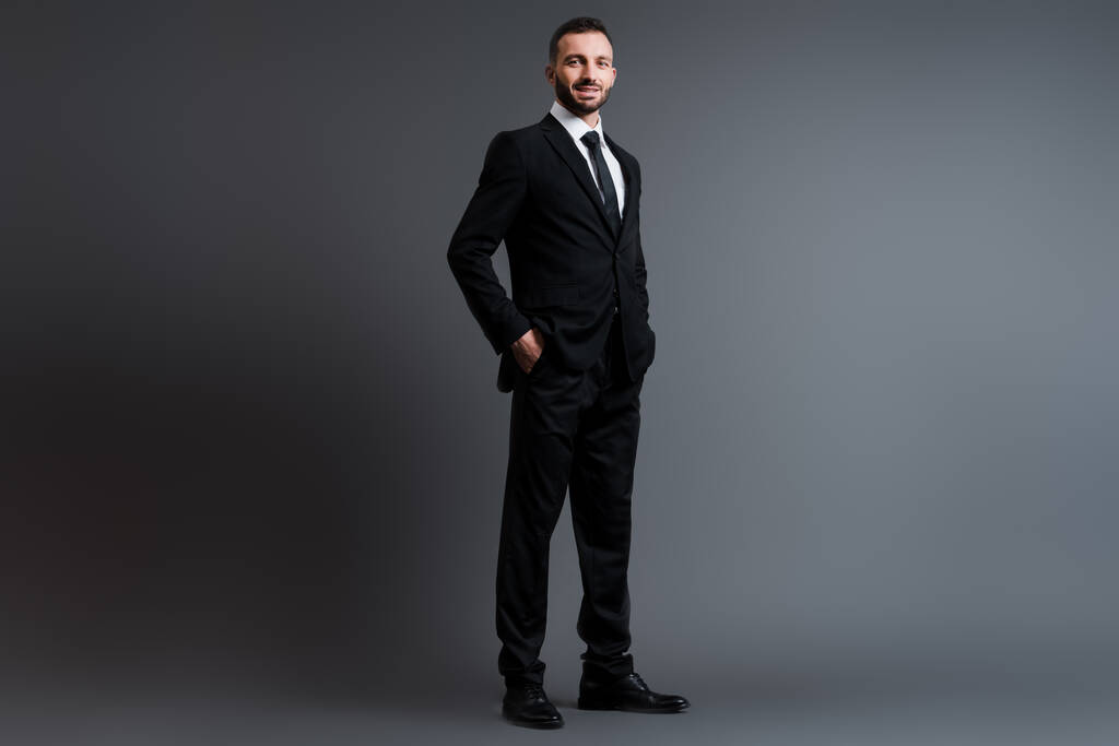 glücklicher Geschäftsmann im Anzug mit Händen in Taschen auf grau  - Foto, Bild