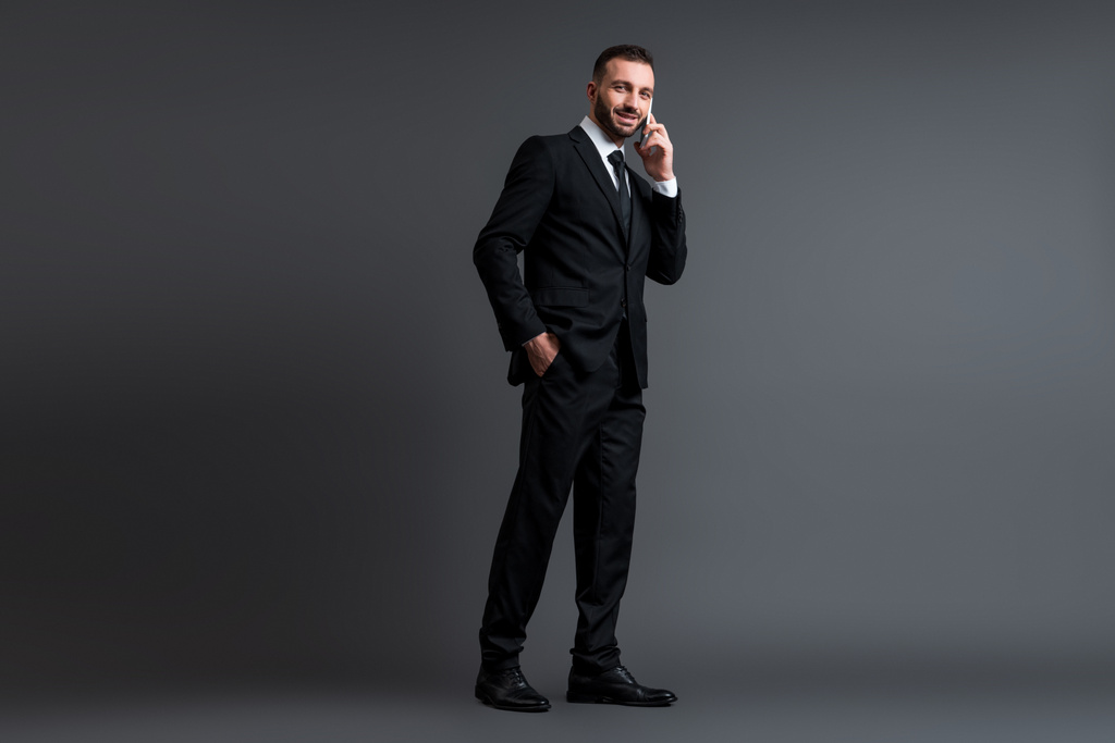 allegro uomo d'affari in giacca e cravatta che parla su smartphone in piedi con la mano in tasca su grigio
  - Foto, immagini