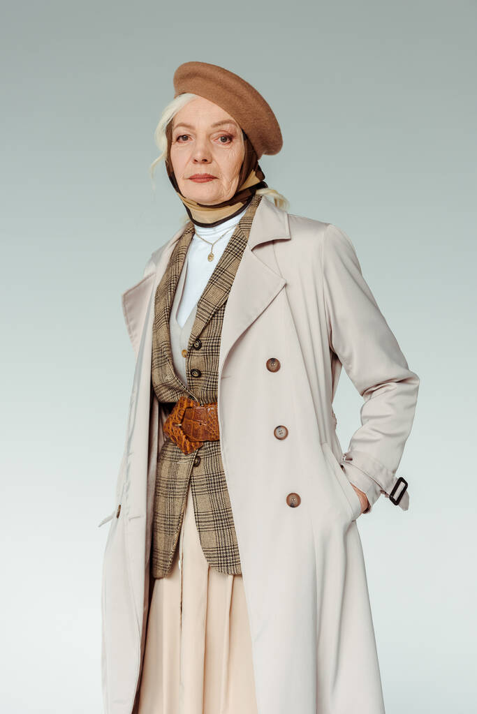 Modieuze oudere vrouw in baret en trench jas op zoek naar camera geïsoleerd op grijs - Foto, afbeelding