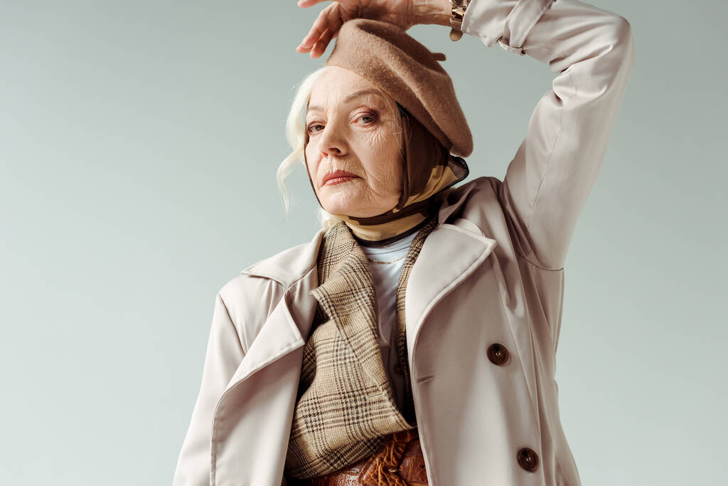 Módní starší žena v trenč kabát při pohledu na kameru při pózování izolované na šedé - Fotografie, Obrázek
