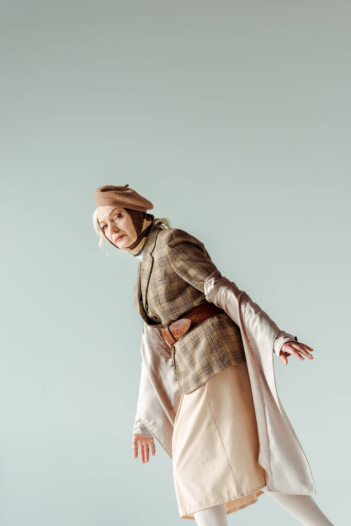 Vue latérale d'une femme âgée élégante vêtue d'un manteau de tranchée et d'un béret regardant une caméra isolée sur - Photo, image