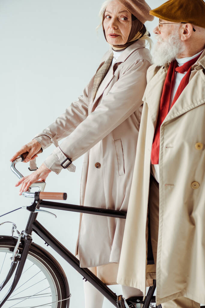 Vista lateral da mulher idosa elegante com bicicleta olhando para o homem elegante em casaco de trincheira isolado no branco
 - Foto, Imagem
