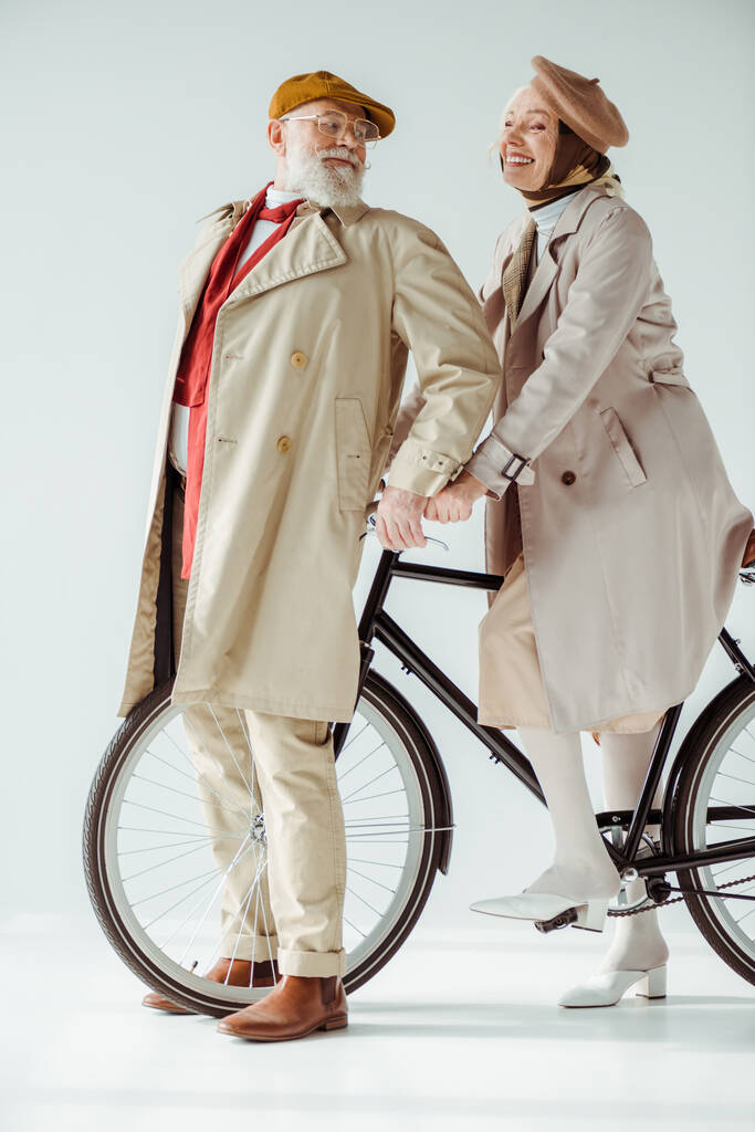 Full length of elegant senior couple with bicycle smiling at each other on white background - Photo, Image