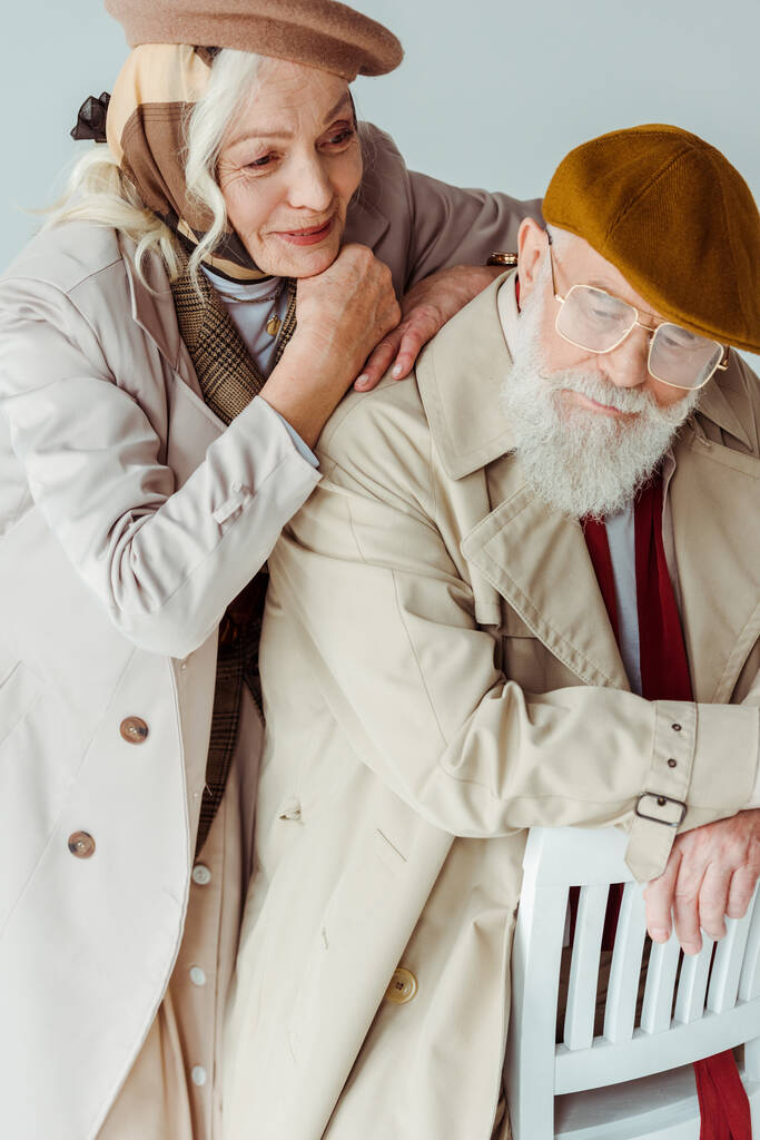 Χαμογελαστή ηλικιωμένη γυναίκα αγκαλιάζει κομψό άνδρα σε καρέκλα απομονωμένη σε γκρι - Φωτογραφία, εικόνα