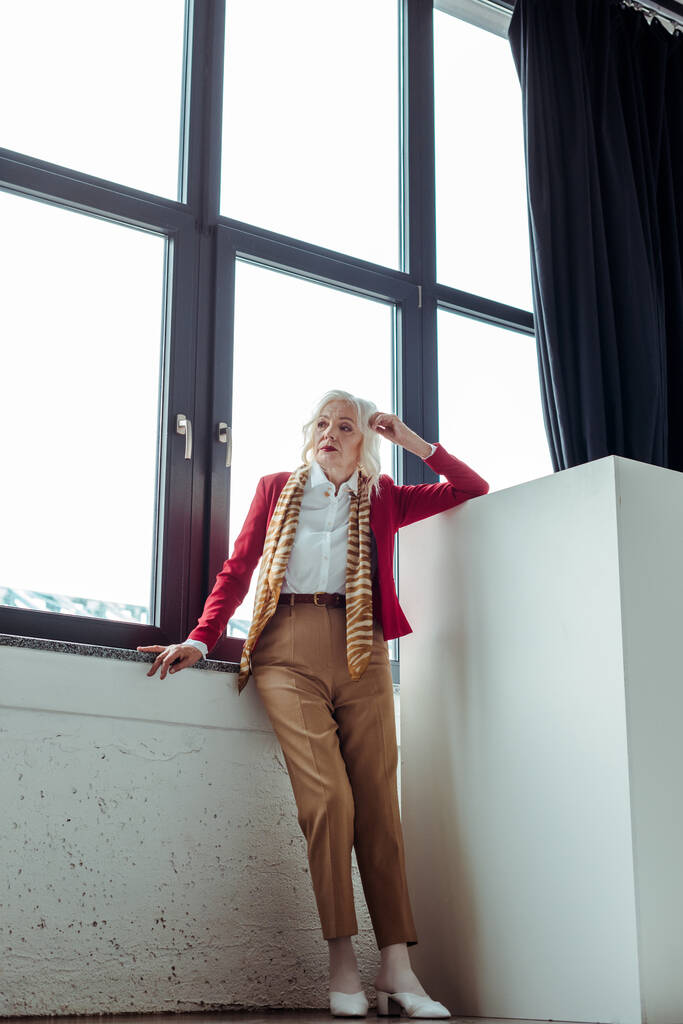 Πλήρες μήκος της κομψής ηλικιωμένης γυναίκας που στέκεται κοντά στο παράθυρο στο στούντιο φωτογραφιών - Φωτογραφία, εικόνα