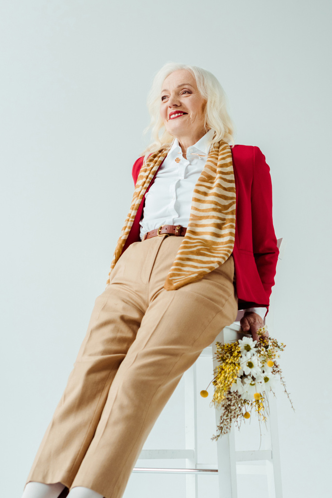 Stilvolle Seniorin hält Wildblumen in der Hand und lächelt auf weißem Stuhl weg - Foto, Bild